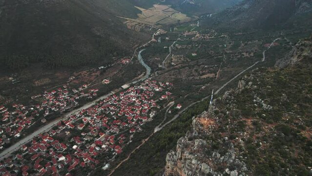 Drone descends above greek flagpole and cliff to reveal town of leonidio at base of valley