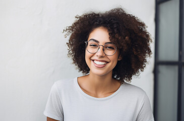 A young biracial of African-European woman in cotton round neck t-shirt with glasses smiling against a white wall near window, concept of aesthetic beauty