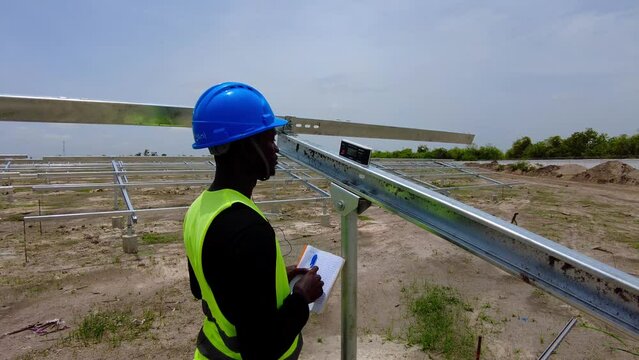 Black African engineer wearing blue helmet taking measurements of solar panel array beam structure tilt angles. Close-up shot