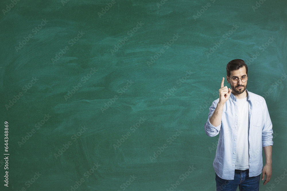 Poster Attractive young european businessman pointing finger up while standing on empty chalkboard background with mock up place. Education and knowledge concept.