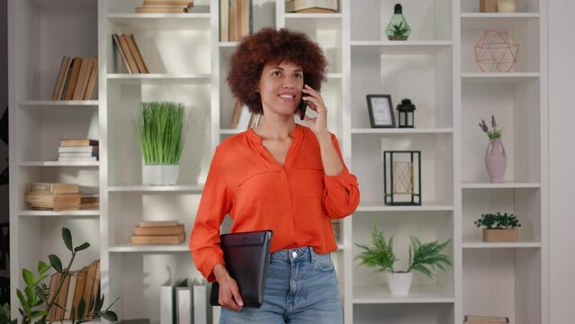 Multiracial young lady holding wireless laptop in case and talking on modern smartphone indoors. Female office worker with curly dark hair smiling cheerfully during mobile conversation at workplace