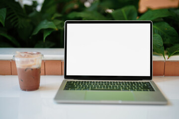 Front view of blank white screen laptop computer with a cup of iced coffee on green wooden table and plant background.