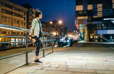 Fototapeta premium Unrecognizable young brunette woman runner with ponytail stretching her legs next to banister before training at night on empty town. Right copy space.