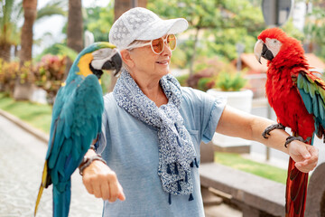 Portrait of mature elderly woman in hat and sunglasses holding on her arms two domestic ara parrots in the park