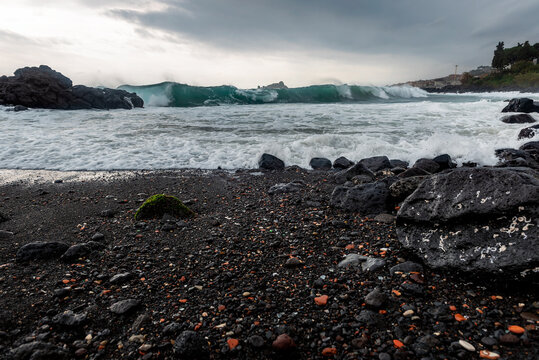 Waves hitting the shore on a cloudy overcast rainy stormy day. Cyclops Coast, black volcanic rocks and send on Sicily, near the town of Acireale