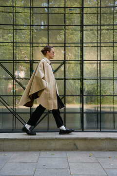 Young androgynous man wearing poncho walking near modern building