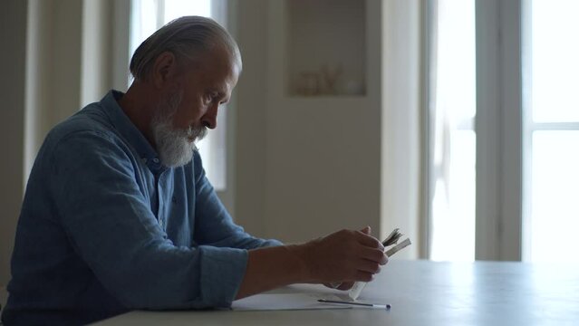 Side View Of Senior Adult Male Writing Financial Information On Paper Counting Counting Russian Ruble Banknotes Sitting At Home Table By Window. Older Pensioner Man Doing Paperwork Planning Budget.