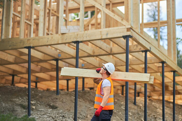 Side view of male builder wearing uniform and helmet, holding wooden boards, gilders, looking up. Man, worker building wooden house in forest in summer. Concept of building.