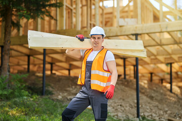 Front view of man, worker wearing uniform and helmet, standing, holding wooden boards, gilders. Builder constructing wooden house, looking at camera. Concept of building.