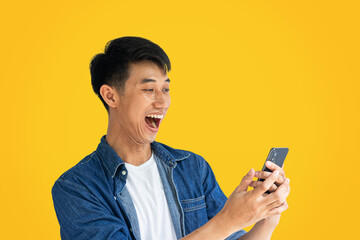 A young Asian man wearing a blue jean shirt is dancing in surprise from a notification on his cell phone. A man is surprised by a message received on his smartphone. Concepts of happiness from online