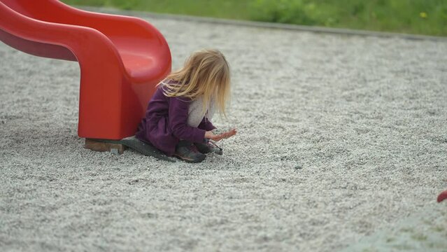 A blonde-haired girl sits near the slide on the modern outdoor playground and plays with stones. Slow-motion.