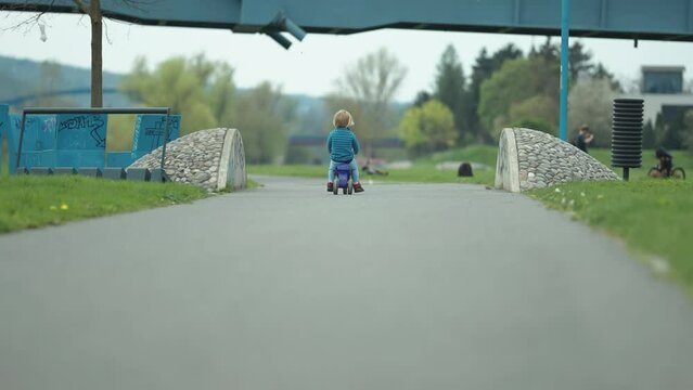 A fair-haired toddler boy rides a kickback scooter along the asphalt path in the park.
