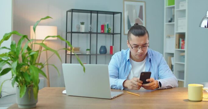Asian male professional holding modern smartphone texting message in home office. Young businessman using helpful mobile apps for business time management organization concept sitting at work desk