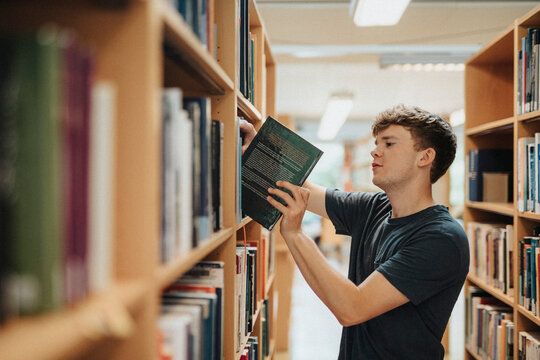 Side view of male student removing book from bookshelf in library at university