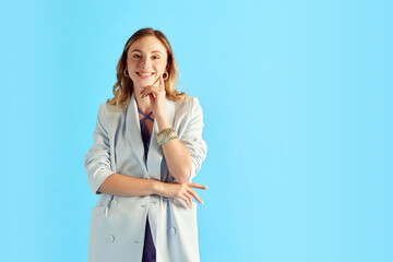 Portrait of beautiful young smiling woman in jacket, wearing stylish accessories, posing against blue studio background. Concept of human emotions, youth, fashion, lifestyle. Copy space for ad