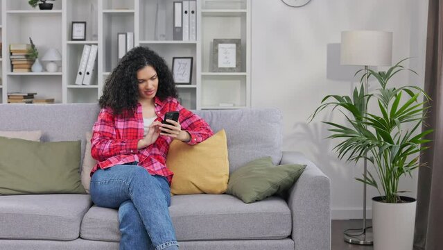 African american young female leaning with hand on soft yellow cushion and surfing internet on smartphone. Casually dressed curly lady visiting online store with clothes while having rest on sofa.