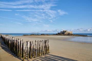 Wooden posts stuck in the sand of a beach and on the horizon an ancient fortress surrounded by the sea with a blue sky with clouds