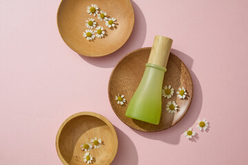 Top view, flat lay of a glass bottle with chamomilla flowers decorated on wooden trays on a pink background. Unlabeled for design packaging. Mockup for advertising