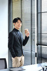 Handsome young businessman holding disposable cup standing in office and looking at camera