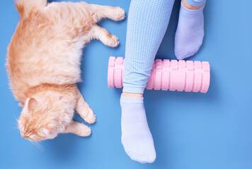 Female legs on foam roller and ginger cat on blue background top view.