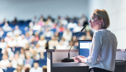 Female speaker giving a talk on corporate business conference. Unrecognizable people in audience at conference hall. Business and Entrepreneurship event