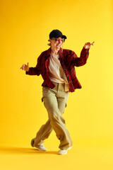 Full-length image of young woman in stylish clothes dancing and having fun against yellow studio background. Hipster. Concept of human emotions, youth, fashion, lifestyle, ad