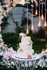 A beautiful large white three-tiered wedding cake decorated with roses stands outdoors in the evening against the backdrop of an arch with electric lamps and garlands. Food photography, holiday.