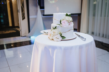 A beautiful delicious two-tiered cream cake decorated with flowers stands on a round table with a tablecloth in a restaurant. Wedding photography, birthday, food.