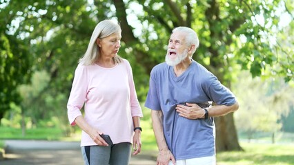 Sick senior gray haired man having a heart attack during morning training in an urban city park. Mature old wife supports her husband and calls for help online through the mobile application on phone