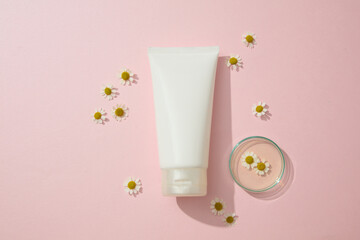 A white plastic cosmetics container for moisturizer cream or facial cleanser displayed on a pink background with chamomilla flowers and petri dish. Top view, space for design packaging