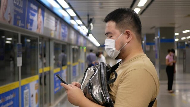 asian young man use 5g smartphone mobile phone in the subway station while waiting for arrival upcoming subway train. Asian man passenger in public transportation with technology
