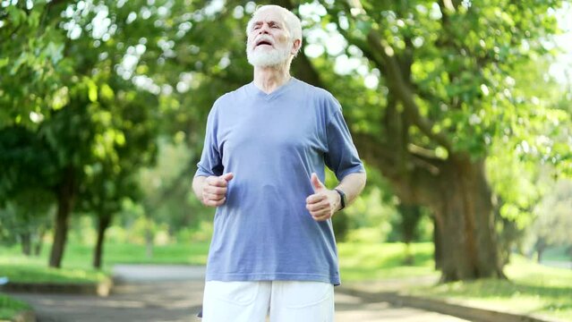 Senior active gray haired bearded man is short of breath during a morning run in an urban city park. Mature old fitness male leans on his knees, wipes the sweat from his forehead and breathes deeply
