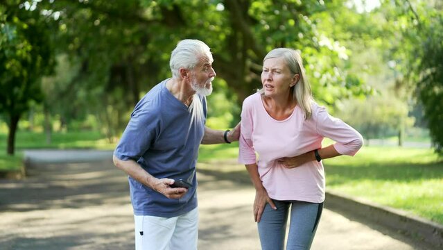 Sick senior gray haired woman having a heart attack during training in urban city park. Mature old husband supports his wife and calls an ambulance by phone. The female needs urgent medical attention