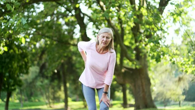 Senior gray haired woman suffering from back pain, sports injury, muscle spasm in urban city park. Sad mature old female having back flank ache and problem after workout outside, massaging a sore spot