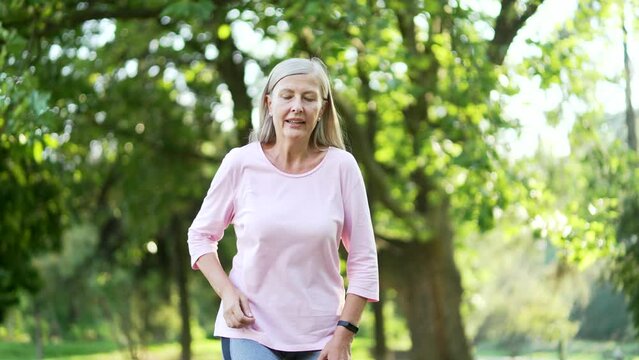 Senior active gray haired woman is short of breath during a morning run in city park. Mature old fitness female leans on her knees, wipes the sweat from her forehead with her hand and breathes deeply