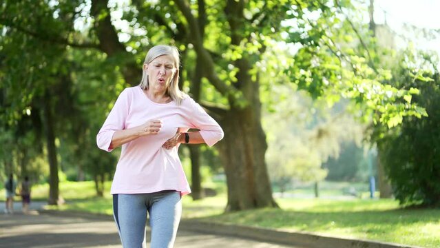Senior adult active gray haired woman has a heart attack while running in an urban city park. Sick mature old female runner suffers from tension in the chest and holds the sore spot with her hand