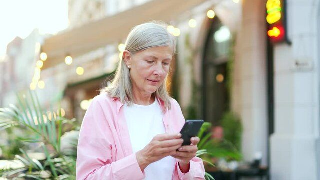 Senior gray haired woman uses a smartphone standing on a city street. Mature old female browsing mobile phone online map application, writes or reads messages, chats online or browses social networks