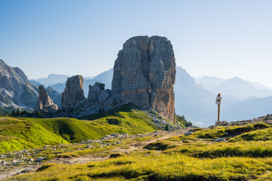 Cinque Torri peaks at summer cloudless morning with green grass lighteen by warm light of sun and guidepost against rocky mountains