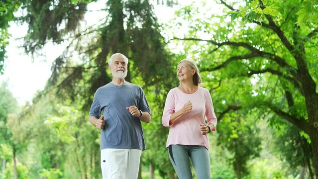 Active married old senior couple jogging in an urban city park. Sporty mature gray haired retired wife and husband enjoys running in nature in morning. Elderly runners during a joint workout in forest