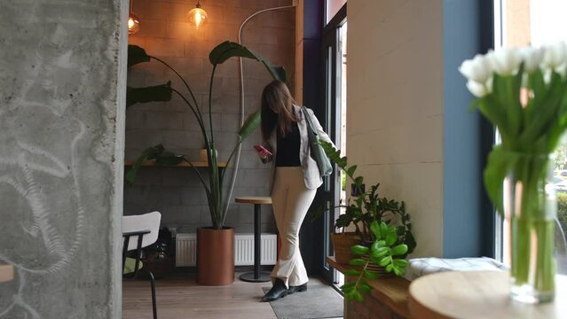 Wide shot interior of cafe entrance with young Caucasian plus-size woman entering in slow motion leaving. Confident charming lady walking checking messages in smartphone app