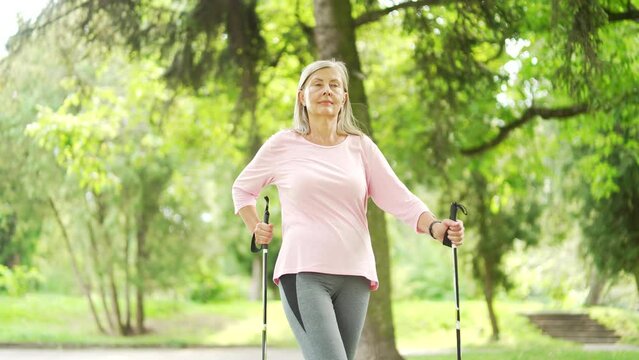 Senior active gray haired female with trekking poles walks in an urban city park. Mature old fitness woman practicing Nordic walking in nature. Elderly sportswoman is engaged in Scandinavian walking