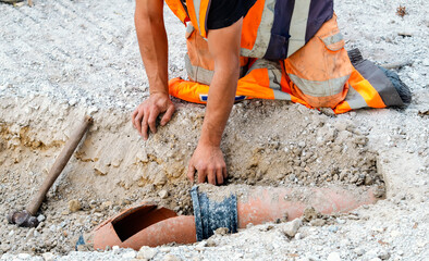 Builder in orange hi-viz clothing hand diggin and exposing damaged drainage pipe for repair