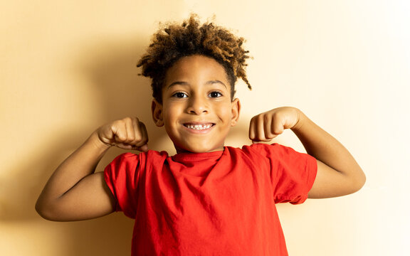 An African boy between 5 and 6 years old with afro hair is happily posing in a brown studio background with a red t-shirt. The little one shows the two biceps with a sign of positivism.