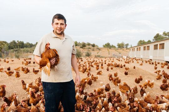 Happy young man standing among flock of hens on poultry farm land