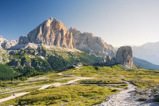 Cinque Torri peaks and Tofane Mountains at summer cloudless morning with green grass lighteen by warm light of sun from right side and path inf front