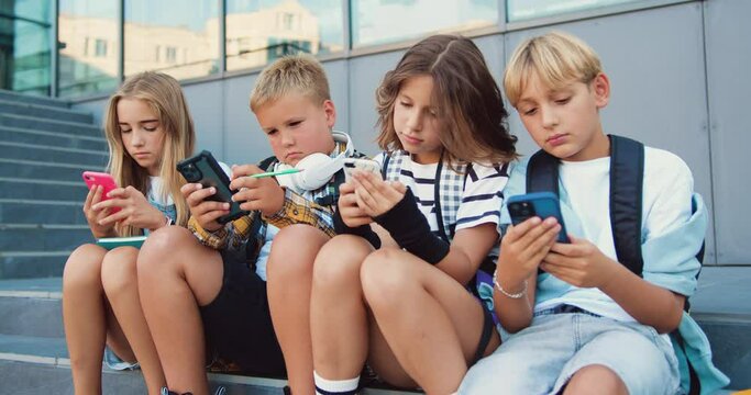 Tenager schoolchildren of two Caucasian girls and boys holding mobile phone and browsing while sitting on stair during school break, oudoors
