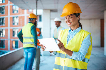 Beautiful mid adult woman architect wearing yellow hardhat at construction site while working on digital tablet. Supervisor wearing safety helmet while working in a building site.  - obrazy, fototapety, plakaty