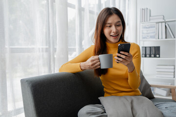 Asian woman is in the living room and uses social media on a smartphone. Female company employee on vacation. Using vacation time and free time to relax at home. Weekend vacation day doing activities.