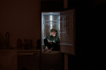 Thoughtful boy with tablet sitting in fridge at night