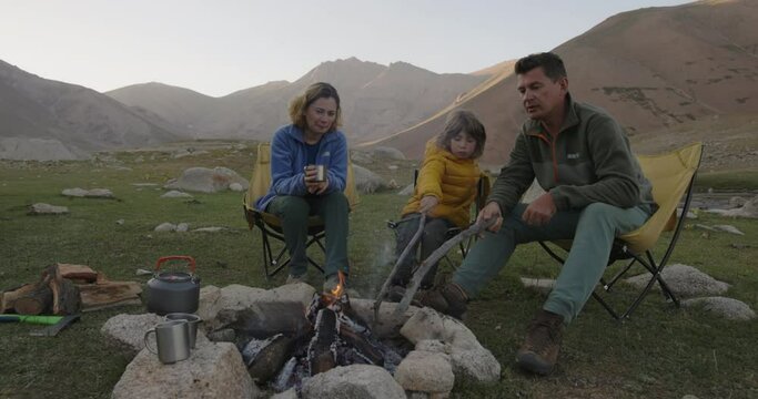 Family by campfire in mountains. Mom dad and child are sitting by fire at sunset. Hike, nature. Family idyll, common hobby. Wellbeing in weekend healthy lifestyle. Joyful peaceful cinematic video